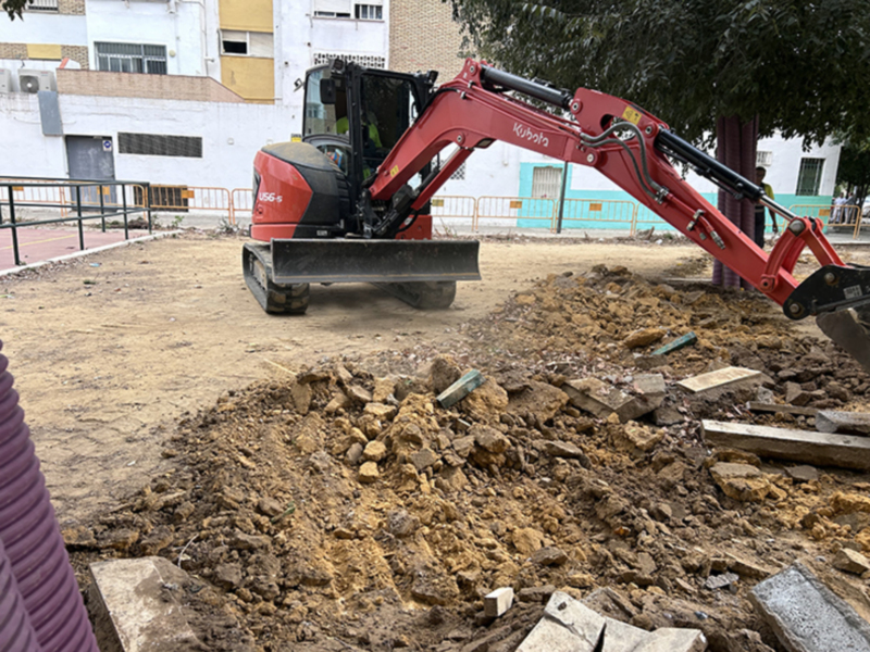 Excavadora roja trabajando en una construcción, con tierra y piedras a su alrededor. En el fondo, edificios blancos con ventanas y una cerca de metal.
