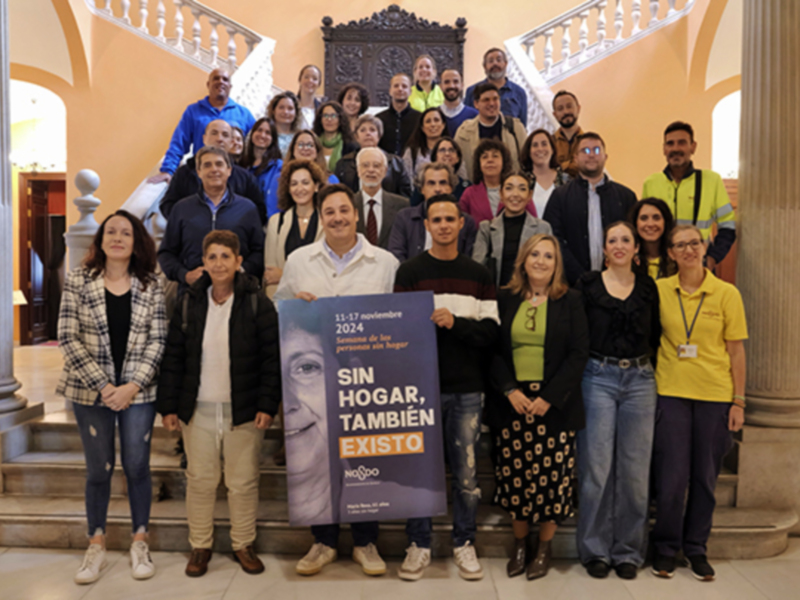 Una imagen de una reunión en un palacio, con personas posando para la foto. En el centro hay una pancarta que dice "Sin hogar, sin futuro" con la fecha 17 de enero de 2024. La imagen muestra a varias personas, incluyendo niños y adultos, vestidos de manera casual. El palacio tiene columnas y una escalera en el fondo.
