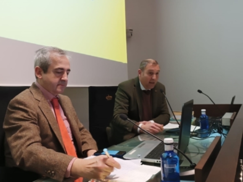 Dos hombres en una sala de conferencias, sentados detrás de un escritorio con micrófonos y laptop, en un ambiente formal.