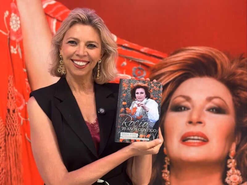 Una mujer sonríe mientras sostiene un libro con la portada de 'Rosa: 20 años contigo' frente a una imagen de la actriz Rosa López, ambos en un fondo rojo.