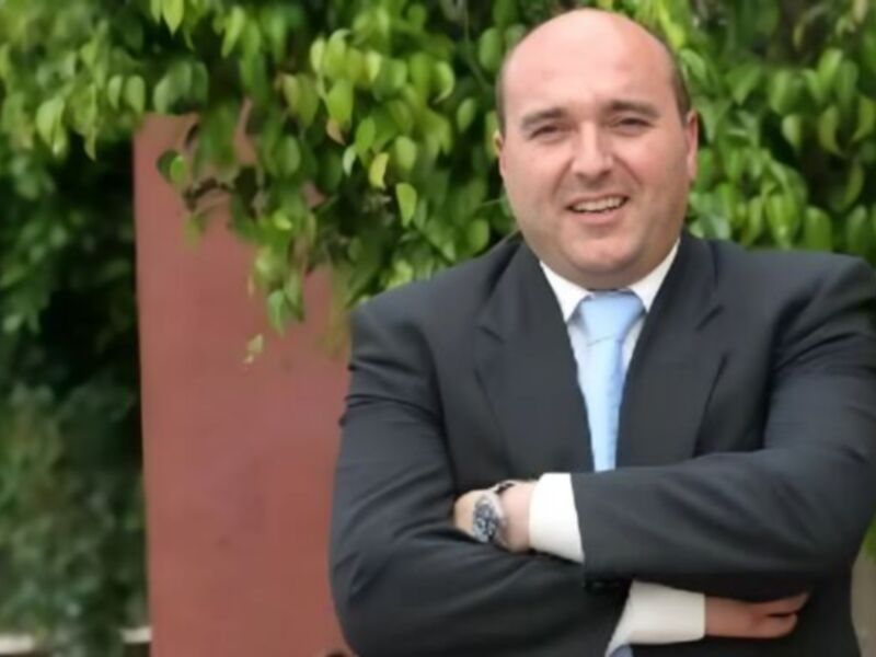 Un hombre en traje de negocios con una corbata azul y brazos cruzados, posando frente a un muro rojo con vegetación en el fondo.