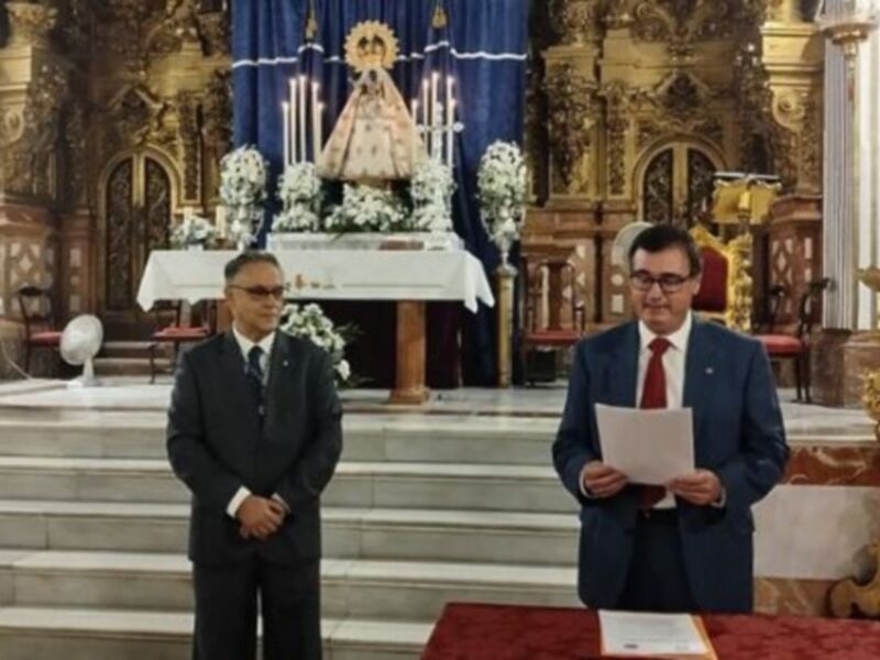 Dos hombres en trajes formales se encuentran frente a un altar dorado en una iglesia, con una imagen de la Virgen María en el centro. La decoración incluye flores y banderas, creando un ambiente religioso y festivo.