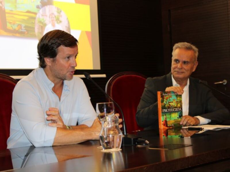 Dos hombres en una conferencia, uno con un libro y otro con un vaso de agua. En el fondo, una pantalla muestra imágenes de personas en un campo. La imagen se toma en un entorno formal con sillas y micrófonos.