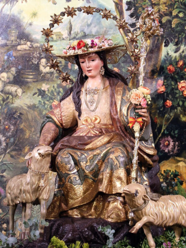 Escultura religiosa de una figura femenina con traje tradicional y flores en la cabeza, rodeada por dos ovejas. Fondo de paisaje con árboles y flores.