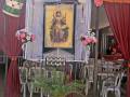 Una decoración festiva con un altar en el centro, rodeado de sillas y flores. La decoración incluye un cartel con una ilustración antropomórfica y elementos tradicionales como un tambor, un sombrero y una caja de música. La decoración es colorida con tonos rojos, amarillos y verdes, y está ambientada en un espacio abierto con una estructura de madera.