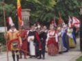 Una imagen de una procesión histórica con personajes vestidos en trajes medievales. Un hombre en armadura y gorro de plata sostiene una bandera española, mientras otros individuos con vestimentas coloridas y sombreros se avanzan por un camino adoquinado. El fondo muestra árboles verdes y una cerca negra, sugiriendo un ambiente rural o de parque.