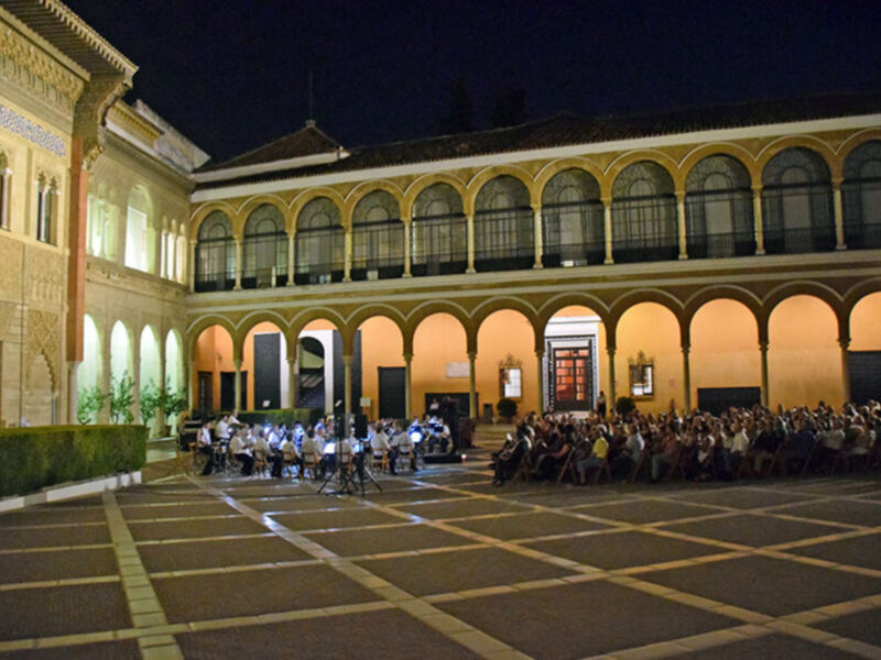 Una escena nocturna en un patio de estilo arquitectónico hispano, con columnas y arcos dorados. Un grupo de personas se encuentra sentado en el suelo, probablemente escuchando a una orquesta que toca al fondo. La iluminación es suave, enfocándose en el patio y las personas sentadas, creando un ambiente sereno y acogedor.