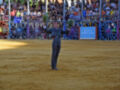 Un hombre en traje negro y corbata sostiene una bandera azul en un escenario de arena, con una multitud de espectadores detrás. En el fondo se observa un cartel azul que dice "Festival Mundial de la Música".