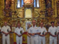 Un grupo de oficiales militares en uniforme blanco se encuentra frente a un altar dorado elaborado, con figuras religiosas y símbolos. El fondo muestra una gran estatua de la Virgen María en el centro, rodeada por otros santos y símbolos religiosos. La escena transmite una atmósfera de respeto y solemnidad, típica de un evento religioso o militar.