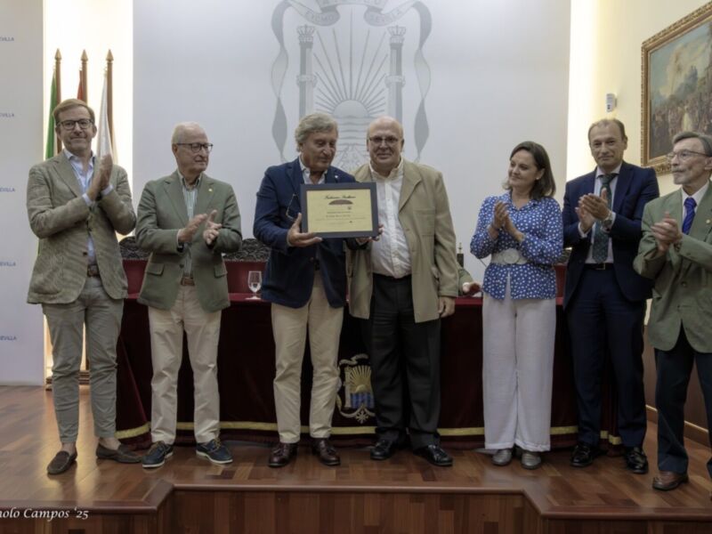 Una imagen de una ceremonia formal donde varios hombres y una mujer se encuentran en un escenario. Algunos están aplaudiendo, mientras que uno sostiene un premio o reconocimiento en una placa. La escena se desarrolla en un entorno con decoración formal y un fondo que incluye una bandera o emblema. La celebración parece ser en un lugar público, posiblemente una sala de conferencias o auditorio.