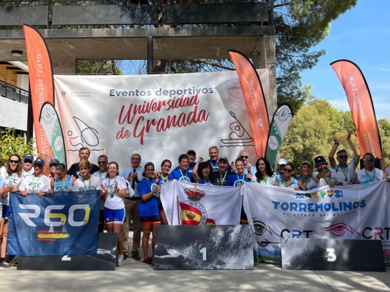 El evento deportivo se lleva a cabo en la Universidad de Granada, con participantes y organizadores posando frente a una pista de atletismo. La imagen muestra un ambiente deportivo y colaborativo, con banderas y logotipos de diferentes instituciones.