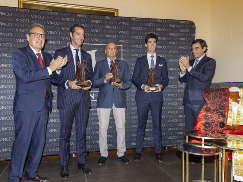 En un evento formal, cuatro hombres en trajes de negocios celebran con aplausos y sonrisas. El fondo es un muro con el logotipo de "VINCI". Una figura en traje y corona se encuentra a la derecha.