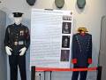 Imagen de uniformes militares expuestos en un museo. En primer plano, una figura de hombre con uniforme y gorra roja, mientras que en segundo plano se puede ver un cartel informativo con fotos de uniformes antiguos. La imagen está ambientada en una sala de exposiciones, con pisos de marmol y paredes blancas.
