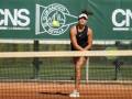 Una jugadora de tenis en acción en un campo de tenis con un fondo verde que dice "Club Náutico Sevilla". La jugadora lleva una camiseta negra con un logotipo de Nike, pantalones cortos y una gorra blanca. La pelota está en movimiento cerca de su raqueta, que es verde con un patrón azul.