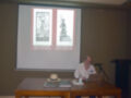 El pié de foto muestra a una persona en un evento de conferencia, posiblemente impartiendo una charla o presentando algún trabajo. En la pantalla de proyección se pueden ver dos tarjetas con el texto "Ex Libris" y "Museo Arqueológico". La persona está sentada en una silla con un sombrero de paja sobre la mesa, y a su lado hay un portátil abierto. La escena parece ser en una sala de conferencias o auditorio, con un fondo neutro y una mesa con algunos materiales.