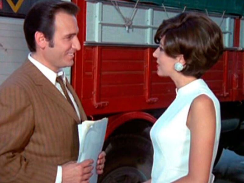 Dos personajes en una escena de la serie "Seinfeld". Un hombre con traje y corbata hablando a una mujer con vestido azul claro. Fondo de un edificio y camión rojo.