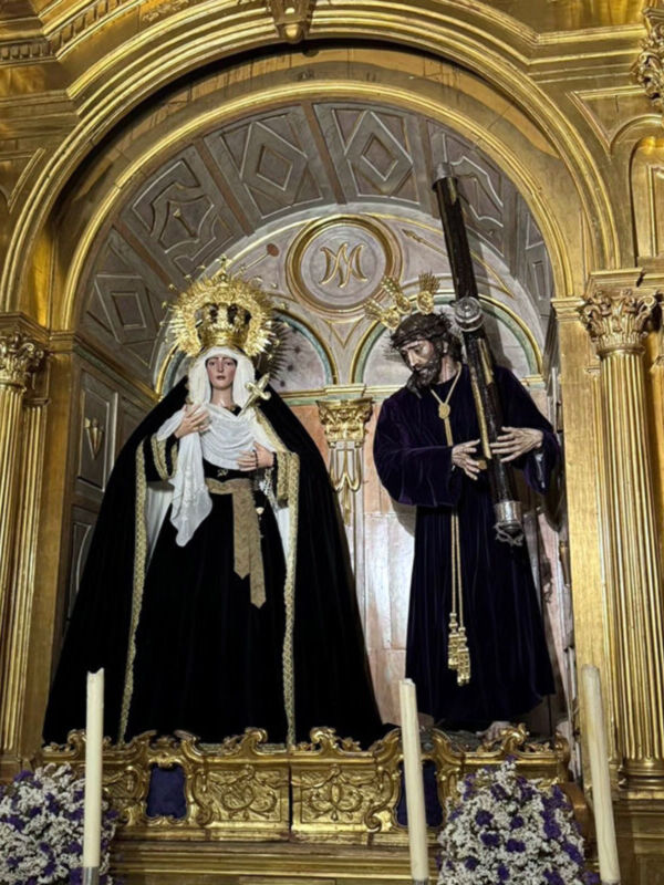 Estas imágenes muestran estatuas religiosas en un altar dorado de una iglesia, con detalles artísticos y decorativos. La estatua principal parece ser una figura femenina con un manto negro y tocado, mientras que la otra estatua parece ser una figura masculina con un manto negro y tocado. El altar está adornado con flores blancas y azules, y hay velas encendidas en el frente. La arquitectura del edificio es de estilo barroco, con columnas doradas y detalles geométricos en el techo.