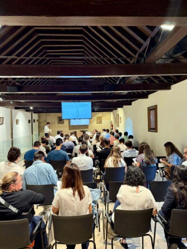 Una sala de conferencias con público sentado en sillas. La pantalla grande muestra información, y hay una persona al frente hablando. El techo tiene vigas de madera y la pared está decorada con cuadros.